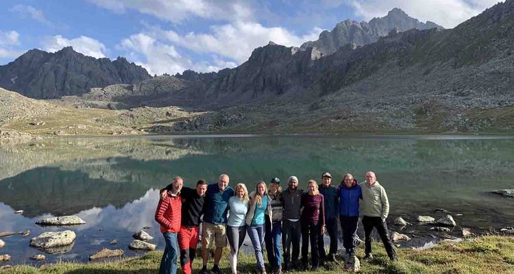 Large trekking group poses beside a high alpine lake with jagged peaks lining the horizon.