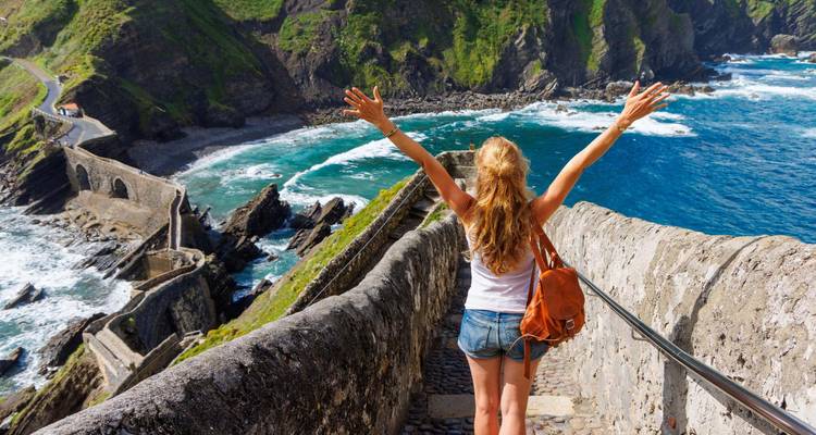 Jeune femme aux bras levés sur un escalier de pierre surplombant une côte sauvage et une mer turquoise.
