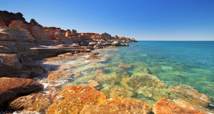 Clear turquoise sea lapping against layered orange and grey rock formations on a remote sunny coastline.