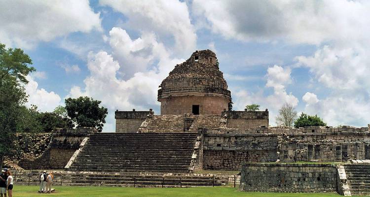 La ruine de l'observatoire en pierre El Caracol à Chichen Itza s'élève au-dessus de larges marches de pierre et d'herbe verte sous un ciel partiellement nuageux.