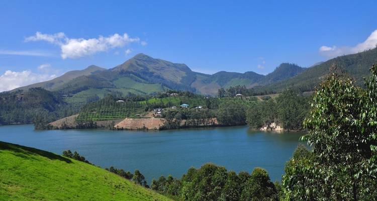 Ein ruhiger See umgeben von sanften, teebedeckten Hügeln und fernen Bergen unter einem klaren blauen Himmel in Kerala.