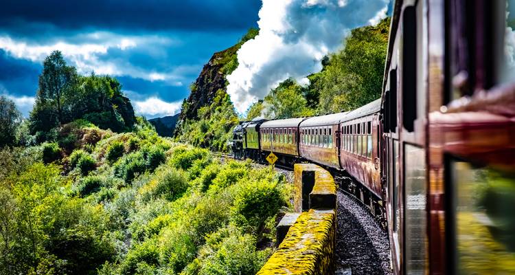 Historic steam train curving through lush green Scottish highlands with dramatic clouds overhead.