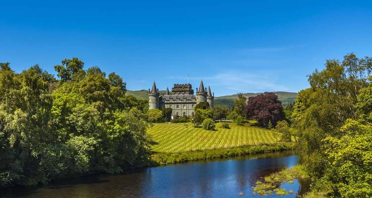 A stone castle surrounded by manicured lawns and dense trees, reflected in a tranquil river.