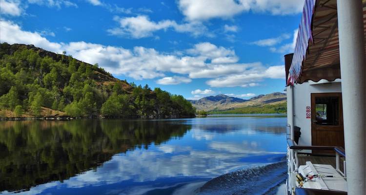 View from a boat gliding across a mirror-like lake bordered by green hills and distant peaks.