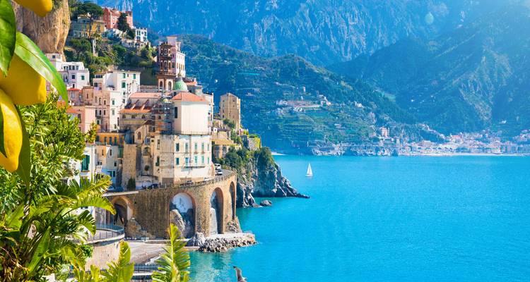 Colourful cliff-top village and terraced hills overlook the turquoise waters of the Amalfi Coast with lemon tree branch framing.
