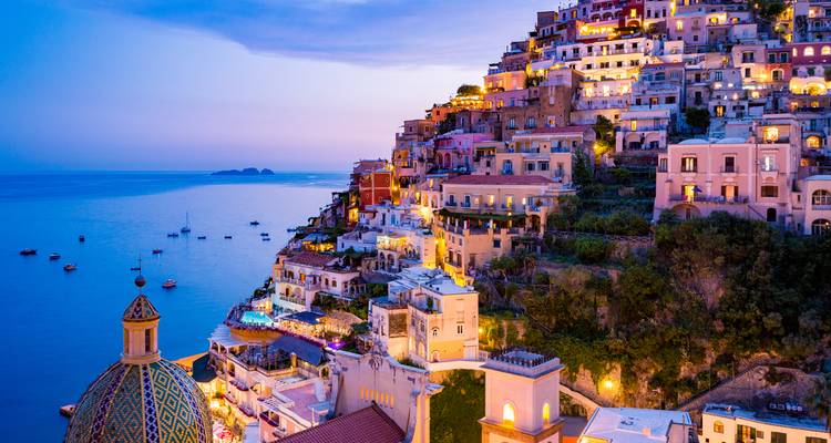 Twilight view of Positano’s pastel houses cascading down cliffs with lights twinkling above calm Mediterranean Sea.