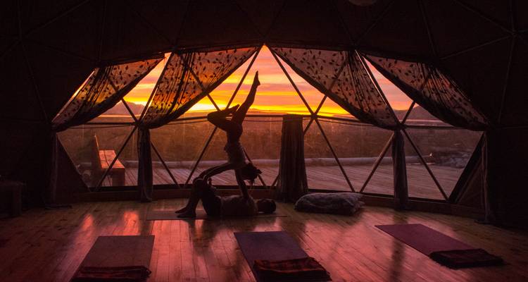 Silhouetted couple practicing acro-yoga inside a geodesic dome at sunset with fiery sky outside.