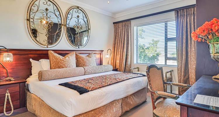 Elegante hotelkamer met groot tweepersoonsbed, sierlijke spiegels en zonlicht dat door een raam naar binnen stroomt.