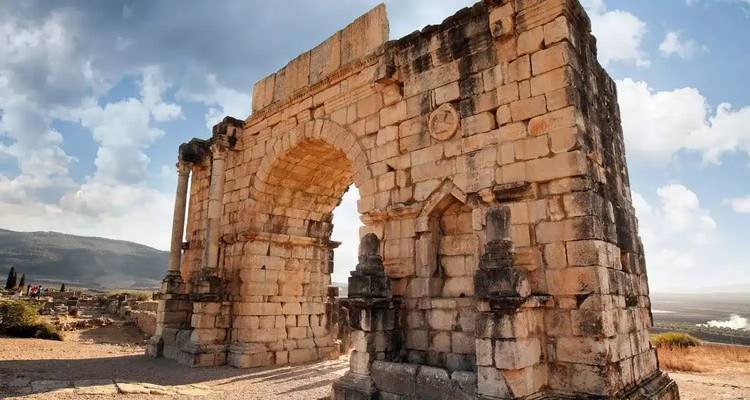 Oude stenen triomfboog staat te midden van verspreide ruïnes in Volubilis tegen een achtergrond van glooiende heuvels en lucht.