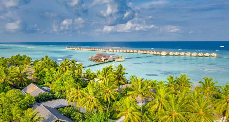 Vue aérienne d'un complexe hôtelier sur une île tropicale avec des rangées de villas sur pilotis entourées d'une mer turquoise et de palmiers luxuriants