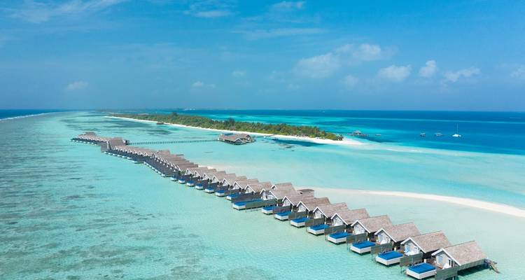 Longue rangée de bungalows sur pilotis au toit de chaume s'étendant au-dessus d'un lagon aux eaux cristallines avec un banc de sable blanc et une île au loin