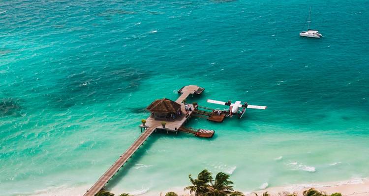 Hydravion amarré à une jetée en bois sur une eau émeraude vive avec un petit voilier à proximité