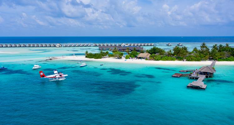Île tropicale avec plage de sable blanc, villas sur pilotis et hydravion amarré au milieu d'un lagon bleu éclatant