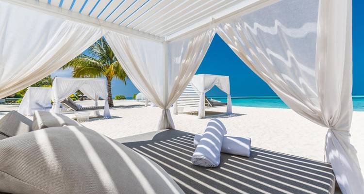 Chaises longues blanches luxueuses avec rideaux sur plage poudrée face à une mer d'un bleu éclatant