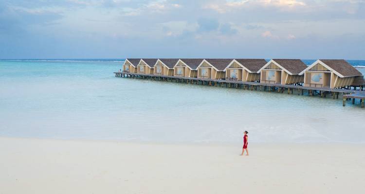 Femme en robe rouge marchant le long d'une plage blanche déserte avec une rangée de villas sur pilotis au-dessus de l'eau en arrière-plan