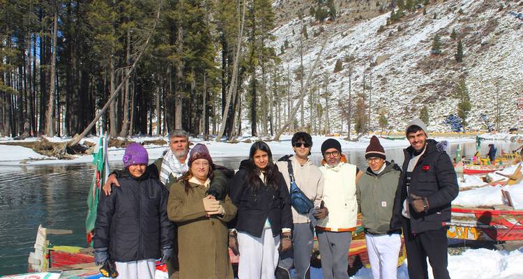Grupo de turistas sonriente posando junto a un lago alpino cristalino bordeado por orillas nevadas y pinos altos en un día soleado de invierno.