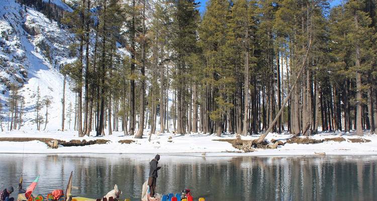 Lago tranquilo bordeado de nieve rodeado por denso bosque de pinos y montañas, con una figura solitaria de pie en un bote colorido.