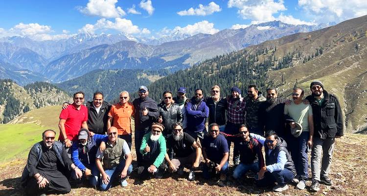 Gran grupo de excursionistas posando en una cresta cubierta de hierba con extensas cordilleras montañosas y cielos azules de fondo.