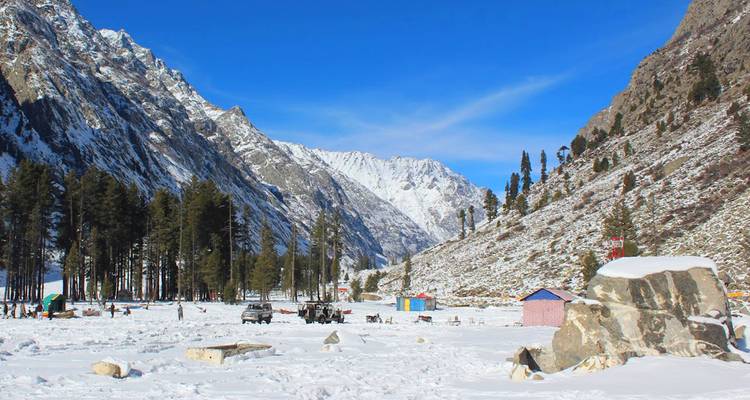 Amplio prado nevado rodeado de picos escarpados y accidentados y árboles coníferos dispersos bajo un cielo invernal azul vívido.