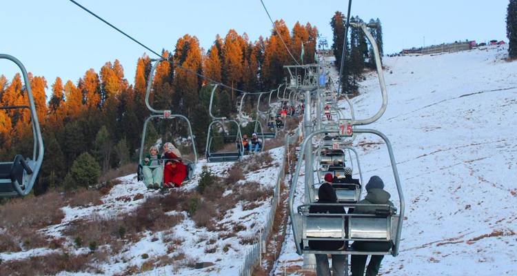 Telesilla transportando esquiadores sobre una pendiente nevada con árboles dorados iluminados por la luz solar del atardecer.