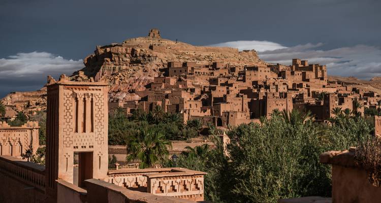 Ancient adobe kasbah village rising on hill under dramatic dark sky, Ait Benhaddou