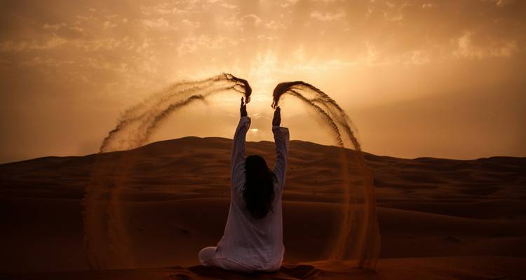 Silhouette of woman sitting on dune tossing arcs of sand against fiery Sahara sunset