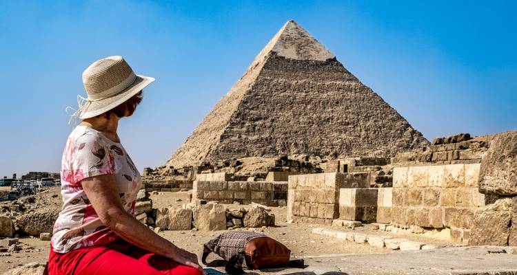 Woman in sunhat sitting and admiring the Great Pyramid of Giza under a blue sky.