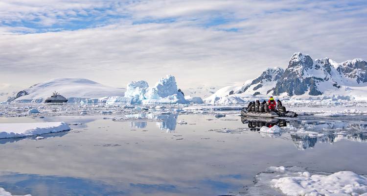 Inflatable zodiac with tourists cruises past icebergs and a cruise ship amid snowy Antarctic peaks reflected in calm water.