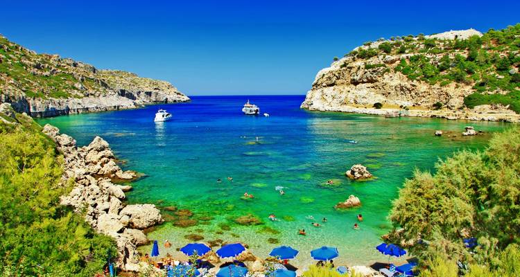 Crique aux eaux cristallines vertes avec des nageurs, des bateaux et des rochers parsemés de parasols sous un ciel bleu profond.