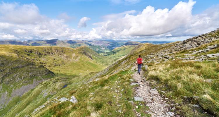 Lone hiker walks a mountain ridge trail with expansive green valley views under a partly cloudy sky.