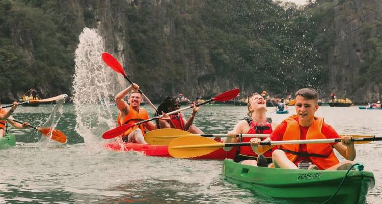 Kayakistas riendo remar a través de aguas esmeralda bajo acantilados de piedra caliza, salpicándose mutuamente con remos brillantes.