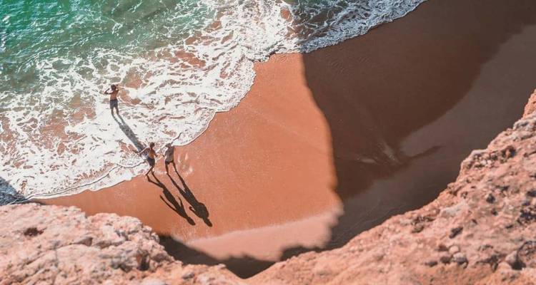 Vista desde arriba de una cala apartada con arena naranja donde dos personas caminan por la orilla espumosa mientras olas turquesas llegan a la playa.