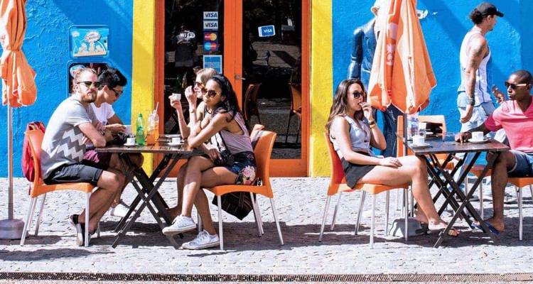 Scène de café en plein air avec plusieurs personnes discutant et buvant à des tables orange devant une façade peinte en bleu vif et jaune.