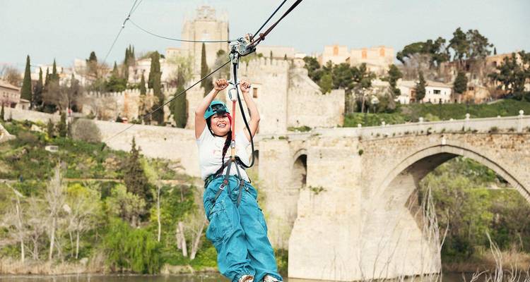 Young woman smiles while zip-lining past a medieval stone bridge and fortified city walls.
