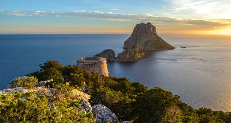 Vue de coucher de soleil de l'île d'Es Vedrà s'élevant des eaux calmes de la Méditerranée avec une ruine de fort côtier au premier plan.