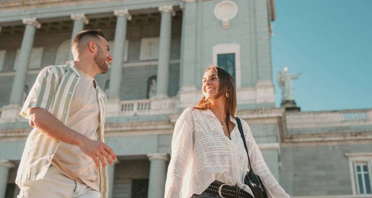 Una joven pareja sonriente camina frente a un magnífico edificio de piedra neoclásico con columnas bajo un cielo azul despejado.