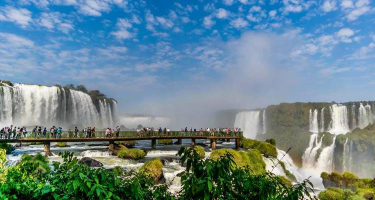 Sweeping view of Iguazu Falls with mist, rainbows and a crowded viewing boardwalk spanning the gorge.
