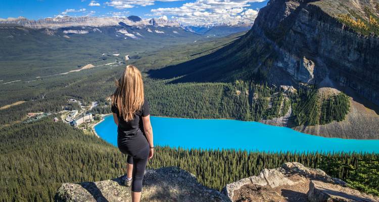 Female hiker stands on rocky ledge overlooking brilliant turquoise Lake Louise and surrounding mountains.
