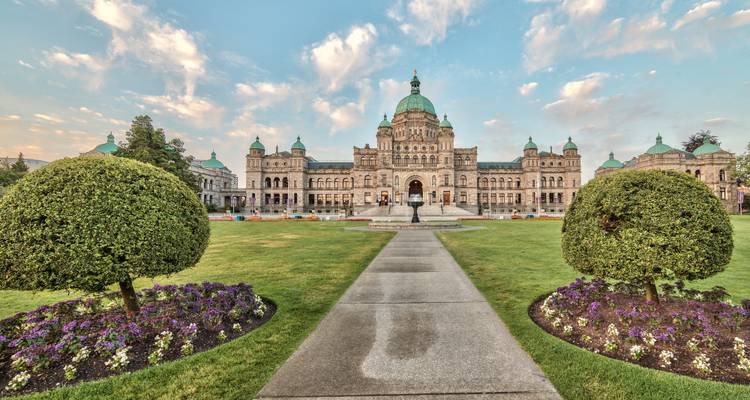 Symmetrical frontal view of the British Columbia Parliament Buildings framed by manicured lawns and flowerbeds at sunrise.