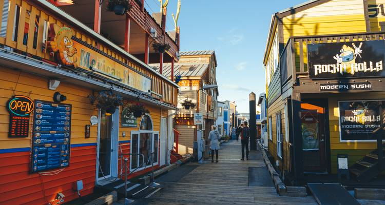 Colorful floating shops and eateries line the wooden boardwalk of Victoria’s Fisherman’s Wharf on a sunny afternoon.