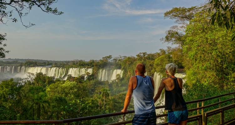 Older couple gazes out over the misty Iguazu Falls from a shaded walkway in late afternoon light.