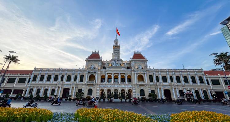 Ho Chi Minh City People’s Committee Building with motorbikes and flowerbeds under blue sky.