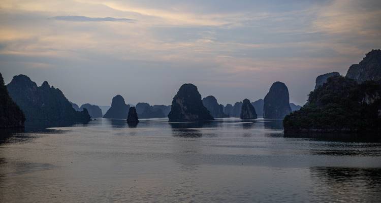 Moody dusk scene of Halong Bay limestone karsts reflecting on still water under pastel sky.