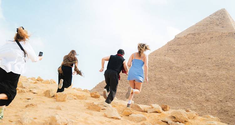 Los viajeros corren por terreno arenoso junto a la Gran Pirámide de Giza bajo un cielo pálido.