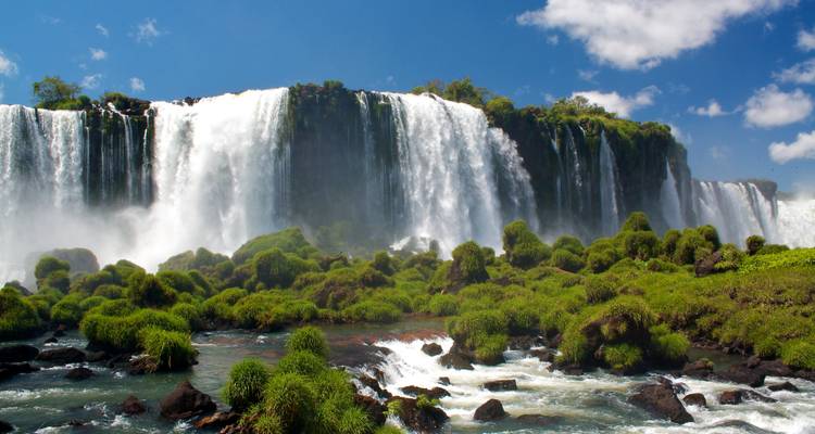 Horizontal sweep of Iguazu Falls spilling over a lush, moss-covered cliff into foaming rapids under a blue sky.