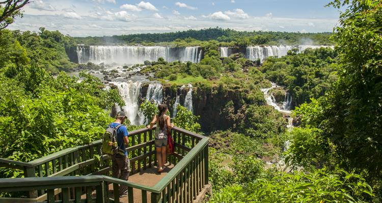 Tourists stand on forested viewing platform overlooking the vast, cascading Iguazu Falls surrounded by lush greenery.