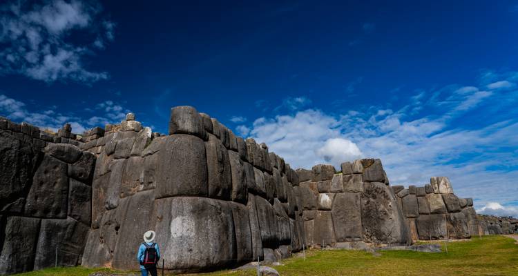Massive stone walls of the ancient Incan fortress Saqsaywaman rise against a vivid blue sky while a lone visitor looks on.
