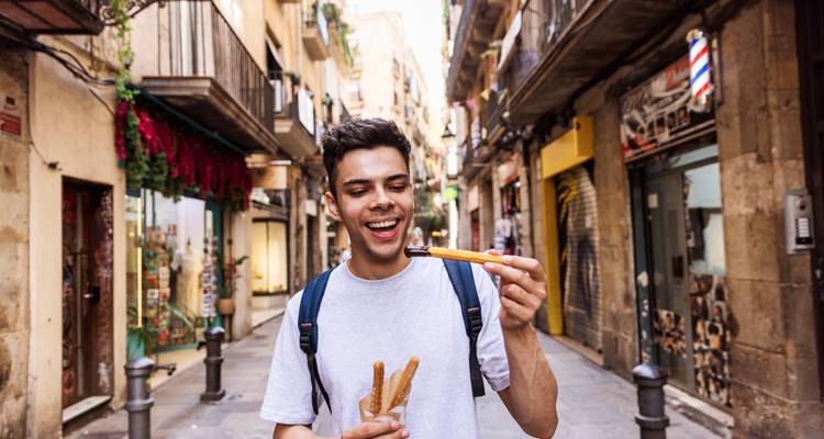 Jeune voyageur souriant tenant des churros dans une rue espagnole étroite bordée de magasins.