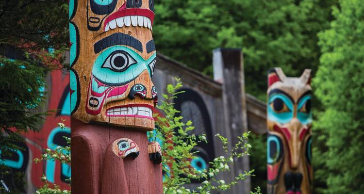 Des mâts totémiques sculptés colorés se dressent parmi une végétation luxuriante dans un décor du Pacifique Nord-Ouest.