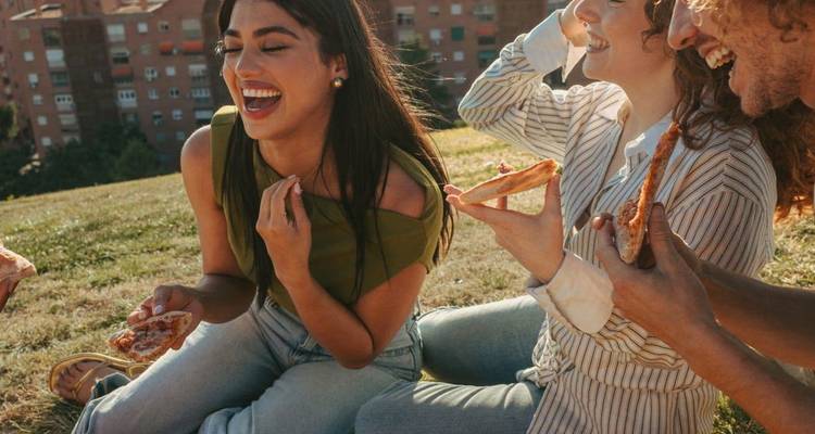 Cheerful friends sharing pizza while lounging on a grassy lawn in the sunshine.
